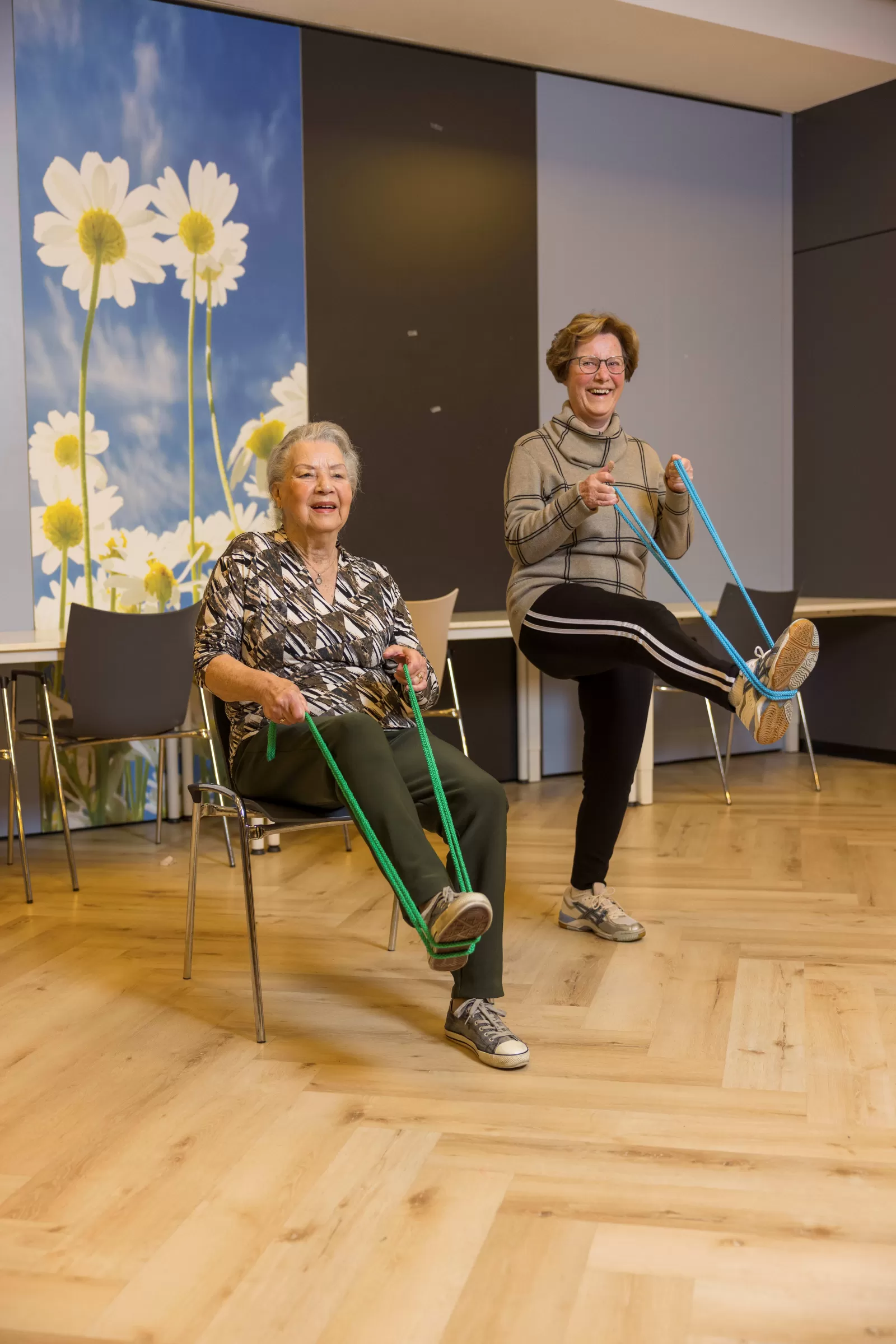 Twee dames tijdens de les Beweeg U Fit. De een doet zittend een oefening, de andere dame doet de oefening staand.